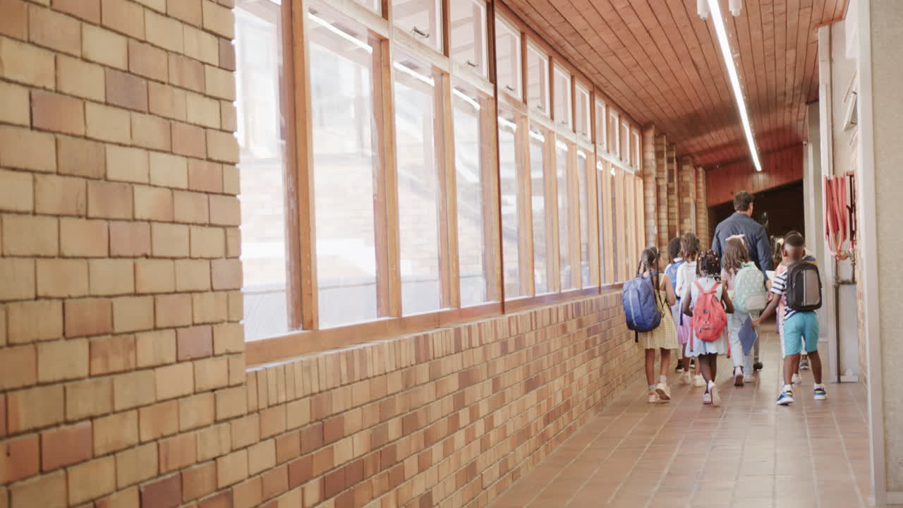 Diverse male teacher with schoolchildren walking in school corridor at elementary school