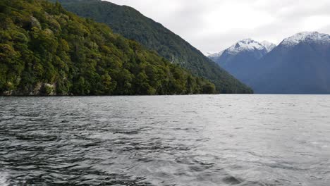 crucero en barco pov en un lago natural rodeado de montañas gigantes con nieve en el parque nacional de fiordland