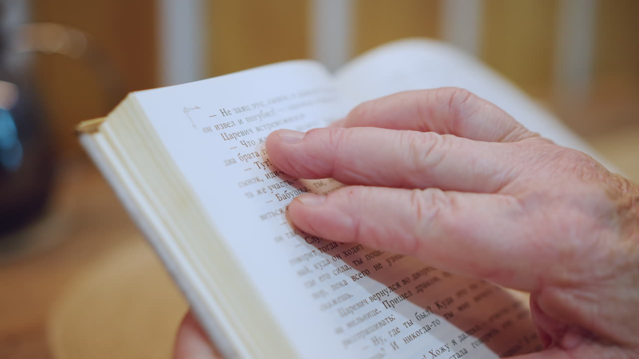 Yakın çekim: Yaşlı eller Braille bir kitabı okuyor, nazik bir parmak kabartmalı noktaları takip ediyor. Arka planda sıcak bir ev masası, yumuşak lamba parıltısı, yaşam deneyimi ve sakin rutini gösteren samimi dokunsal odaklanma.