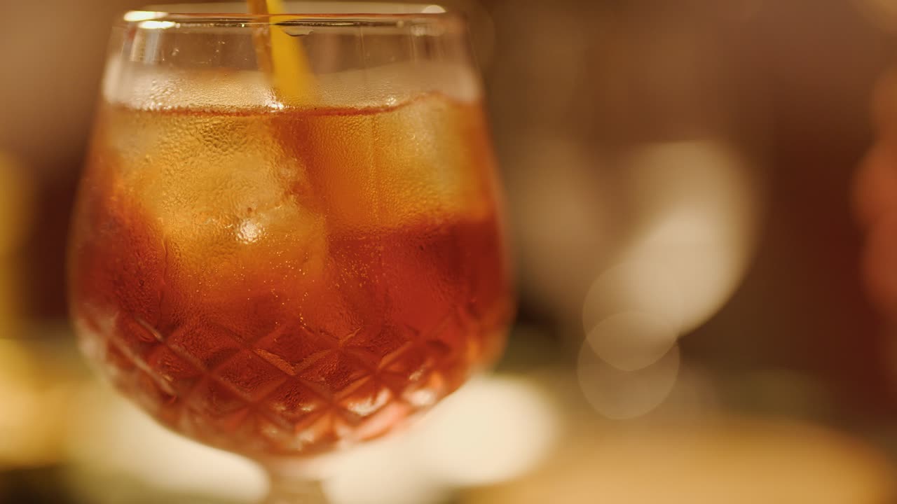 Una mano mescola un cocktail ghiacciato in un bicchiere decorato, illuminazione ambientale calda, messa a fuoco selettiva, sfondo sfocato