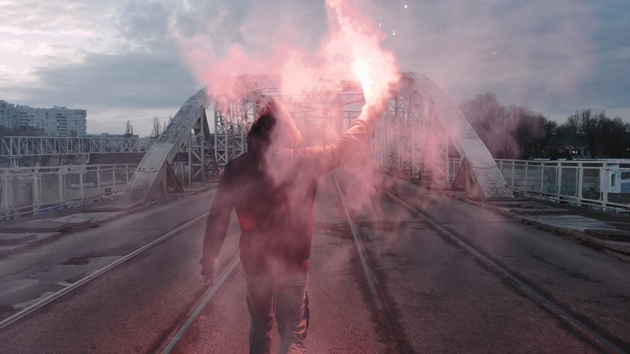 은 강철 프레임 다리 아래의 도로에서 빨간 불타는 신호 플레어와 함께 후디와 발라클라바의 젊은 남자, 느린 움직임