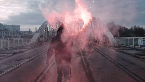 young man in hoodie and balaclava with red burning signal flare on the road under an old steel frame bridge, slow motion