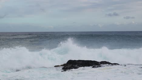 a set of powerful ocean waves hammers a dark coastal rock, sending white spray and foam across the frame, a moody, wind-stirred seascape of raw energy