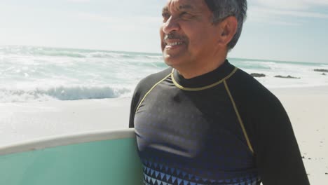 hombre hispano mayor feliz caminando por la playa con una tabla de surf