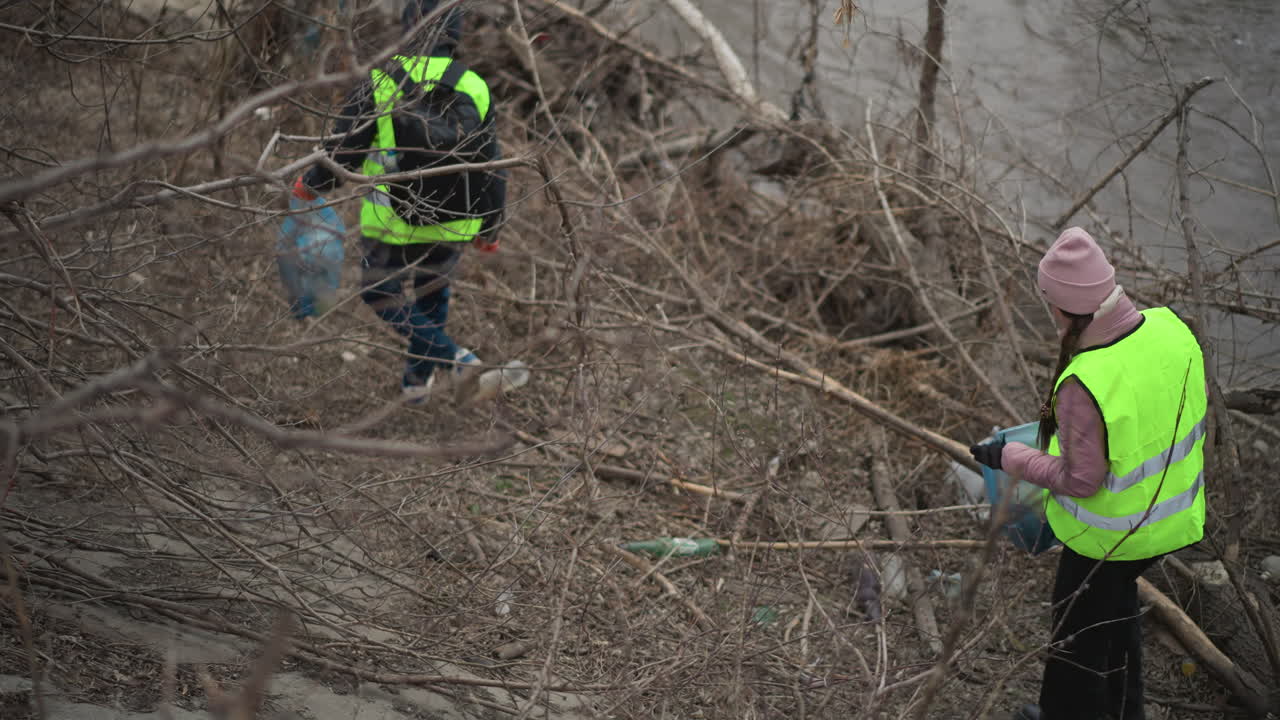 Volunteer in reflective vest and gloves with backpack collecting plastic waste and trash along polluted riverbank surrounded by leafless branches during environmental cleanup effort