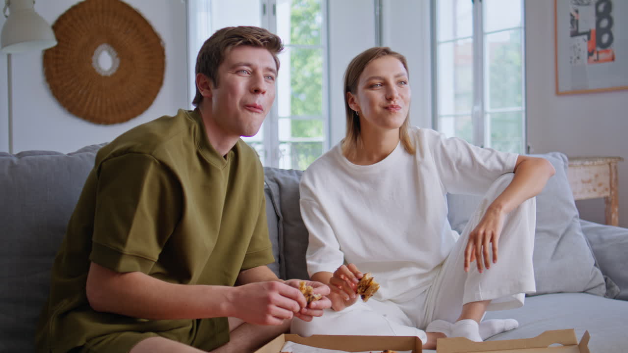 Weekend couple eating pizza sitting sofa in front tv. Happy lovers watching show