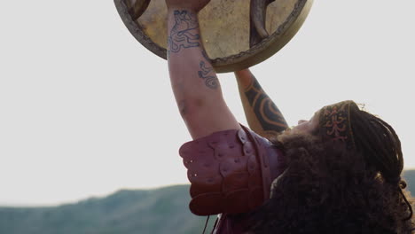 obese man plays shamanic tambourine praying against hills