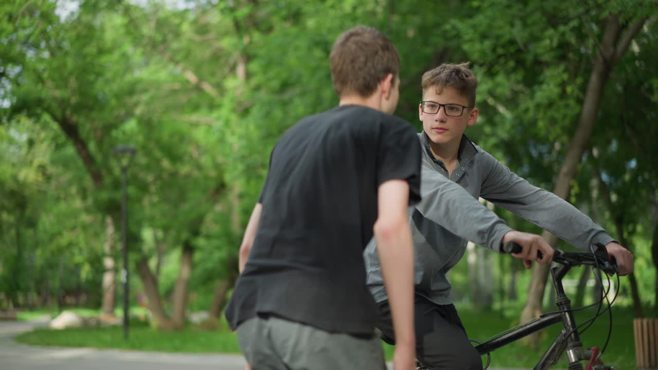 un niño joven con gafas se sienta en su bicicleta, conversando con su amigo con camiseta negra que está sentado, el niño se levantó y se movió hacia él, con vegetación en el fondo