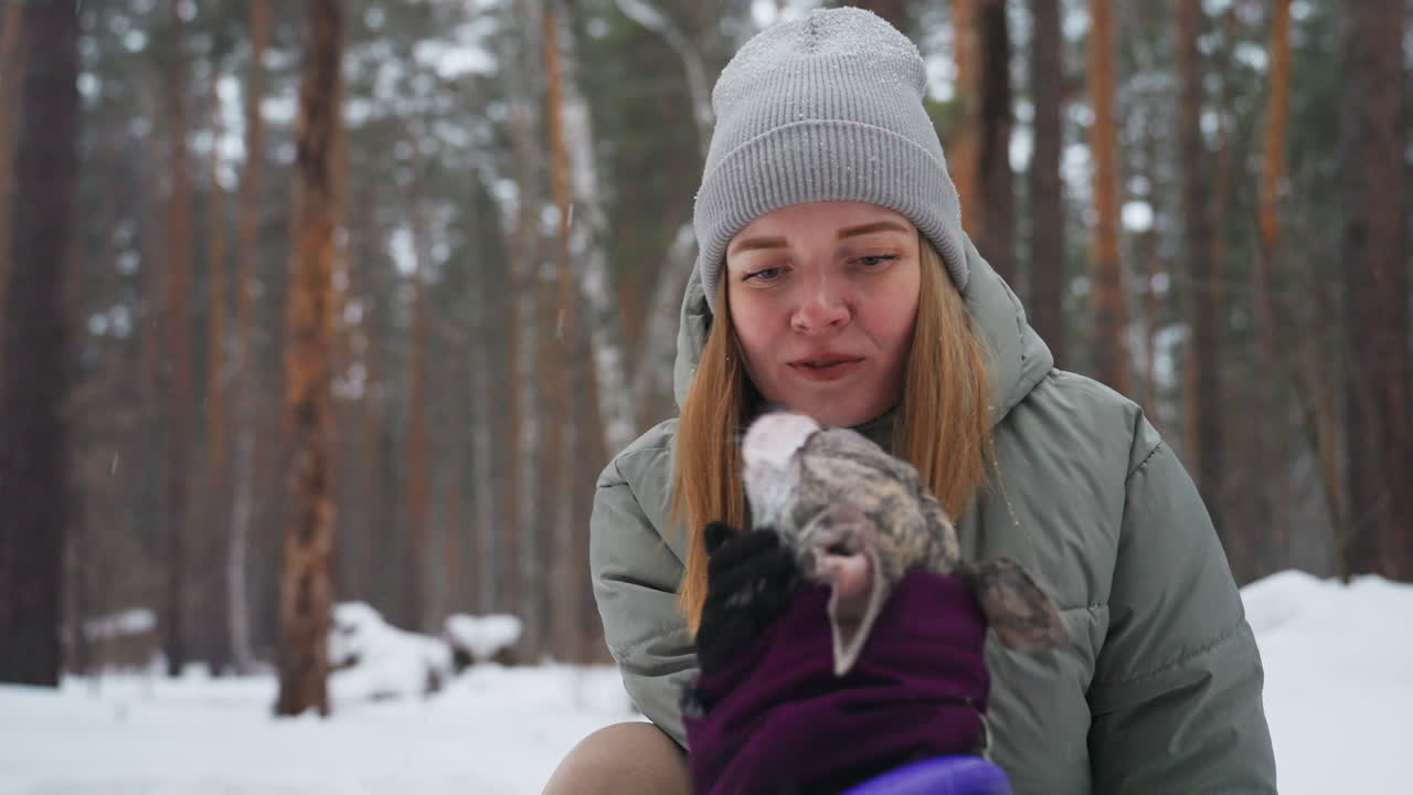 woman in grey puffer jacket wearing knit beanie kneeling in snowy pine forest gently cupping brindle greyhound face in purple coat with gloved hand showing affectionate bond before winter walk
