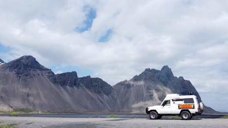 Expedition-campervan-driving-close-by-water-parallel-to-mountains-below-a-white-cloudy-sky