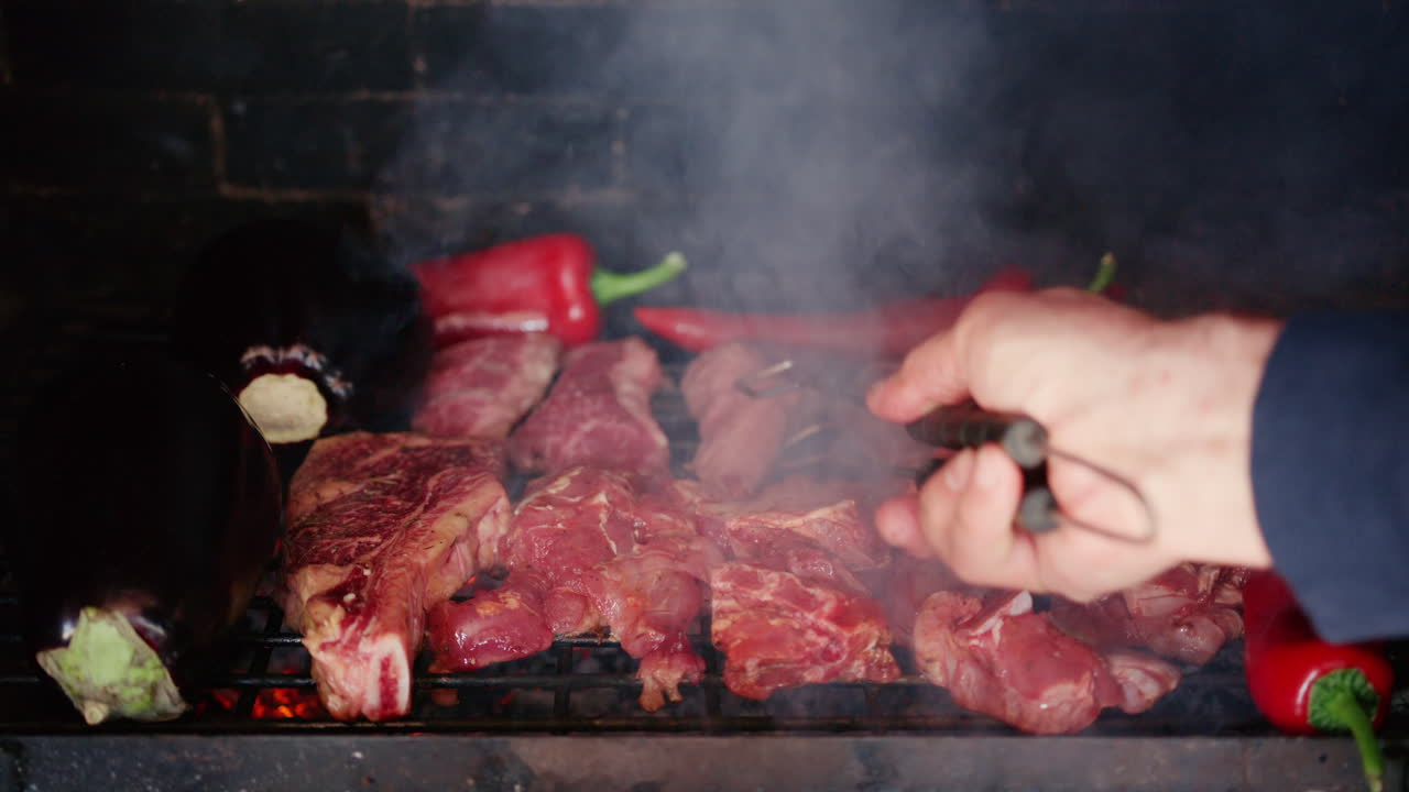 Carne cocinándose en una parrilla con berenjenas y pimientos rojos, con humo elevándose