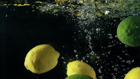 primer plano de limones y limas maduros que caen en agua con gas sobre fondo negro, haciendo un cóctel de cítricos, bebiendo limonada fría, disparando agua carbonatada con frutas flotantes y burbujas