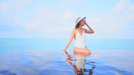 Asiatische-Frau-In-Weißem-Monokini-Und-Skimmer-Sonnenhut,-Die-Am-Rand-Des-Infinity-Pools-Auf-Dem-Dach-Sitzt-Und-Die-Spitze-Des-Hutes-Berührt-Und-Die-Sicht-Verändert,-Vorderansicht-In-Zeitlupe,-Kopierraumvorlage