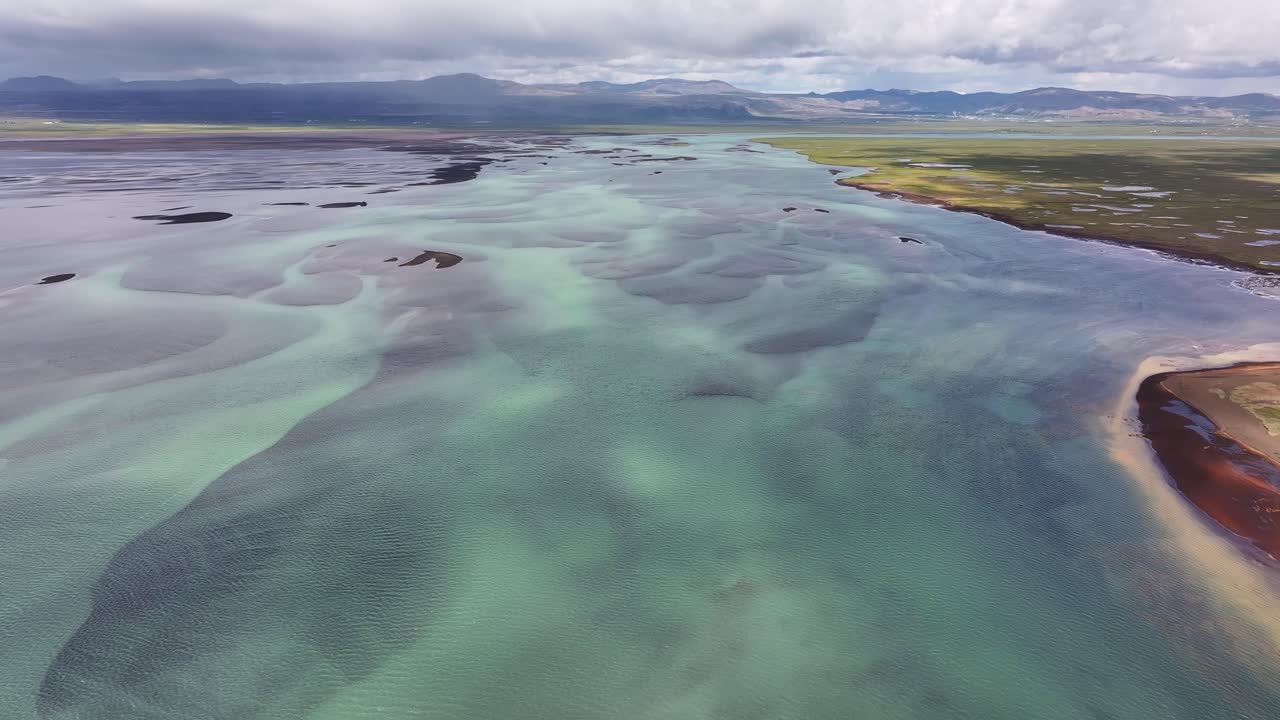 Aerial video pushing back over the gorgeous turquoise water of the Olfusa River in Southern Iceland with mountains off in the distance peaking out before the clouds