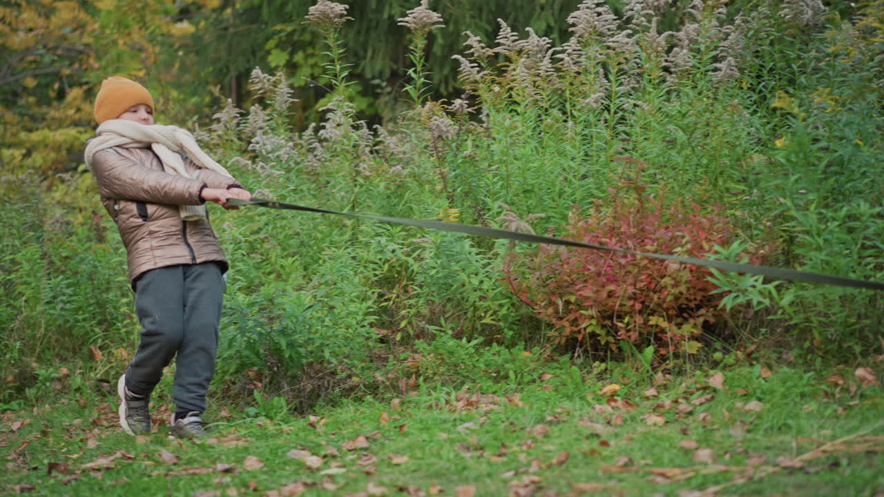 young girl in cozy jacket and knit hat tightly grips leash while being pulled forward by unseen dog or object through vibrant autumn forest trail with scattered leaves and colorful foliage all around