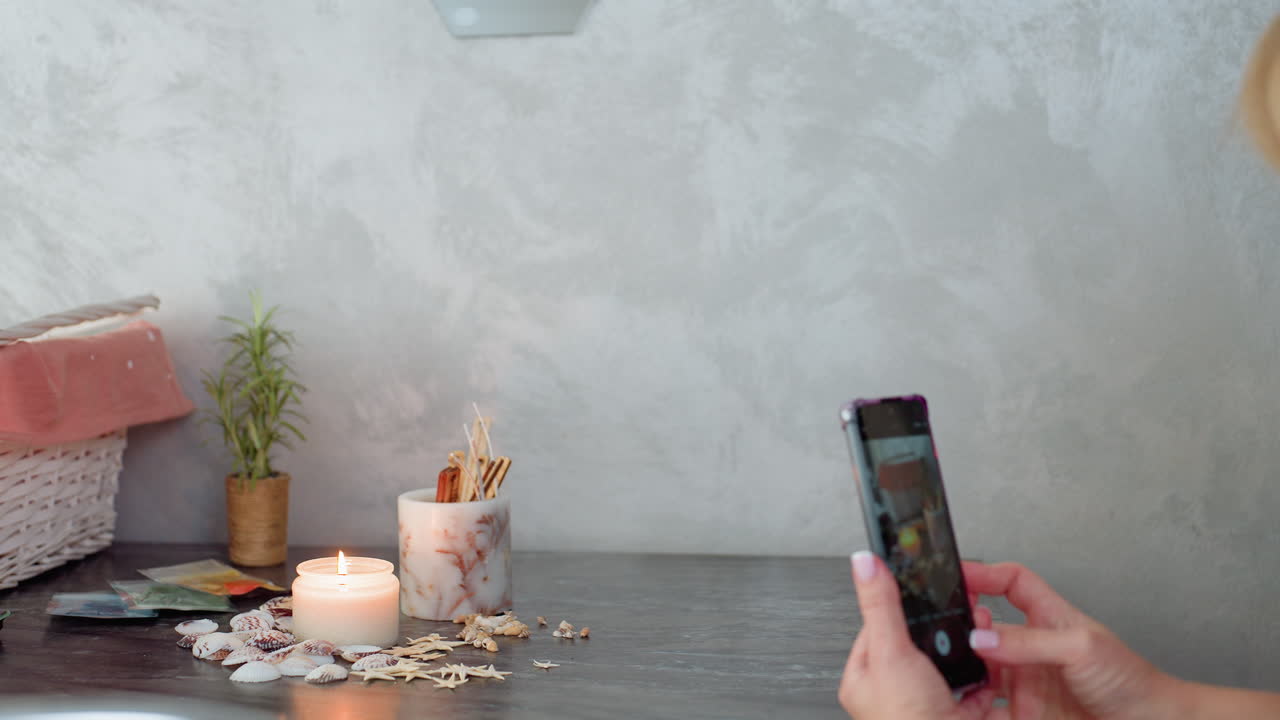 Happy woman wearing bracelet captures cozy candlelight arrangement with smartphone on gray table, surrounded by seashells, small plant, and decorative sticks in soft calm indoor setting
