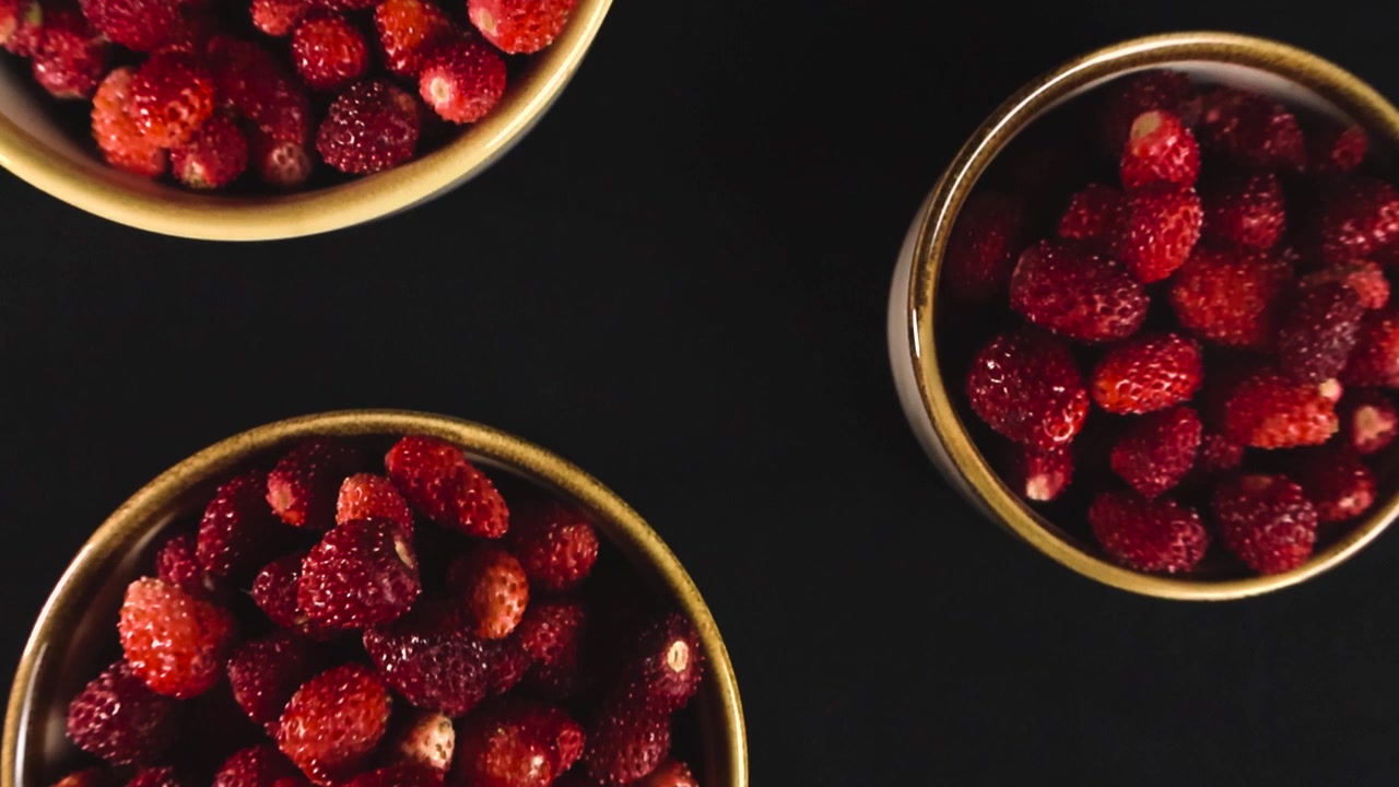 Zwei Keramikschalen voller frisch gepflückter, reifer roter Walderdbeeren, die sich langsam vor schwarzem Hintergrund drehen und ein gesundes, biologisches vegetarisches Sommerdessert aus dem Wald darstellen.