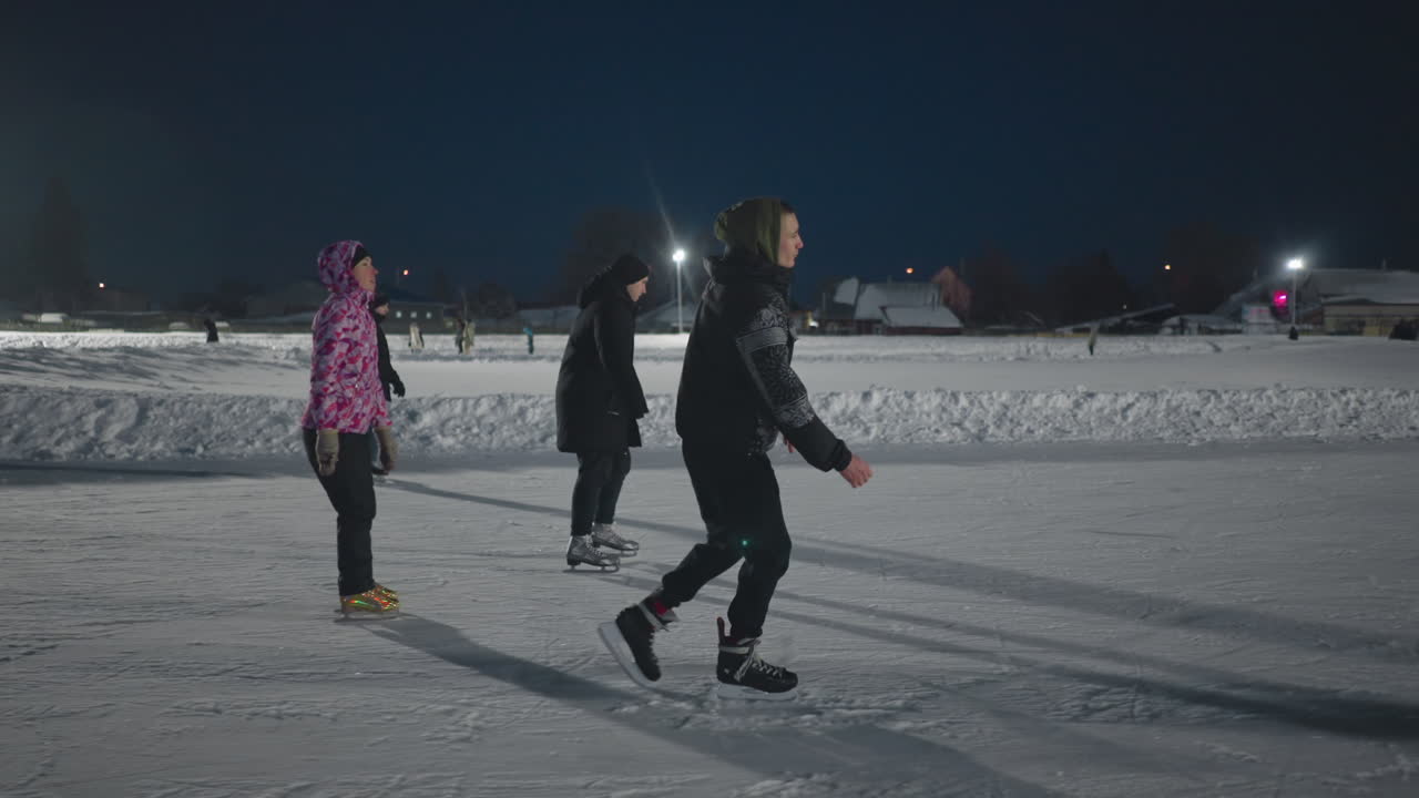 side view of young people gliding on illuminated outdoor ice rink during winter evening surrounded by snowbanks and scattered skaters in distance with relaxed body posture and engaged expressions