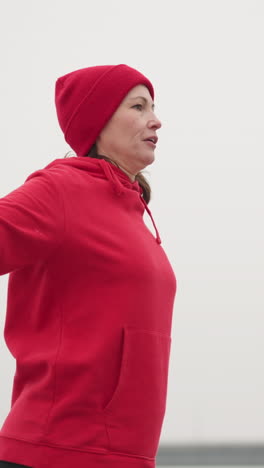 mujer con capucha roja y guantes estirando los brazos al aire libre durante el entrenamiento en un entorno invernal brumoso con postes de luz y características urbanas mínimas en el fondo
