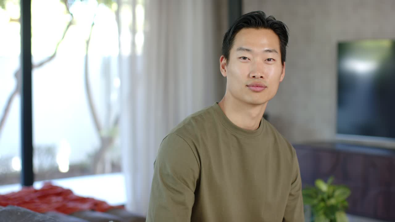 オリーブ色のシャツを着たアジア人男性がソファに座っており、カメラに気づいて目を開け、顔を向け、カメラに微笑んでいる。