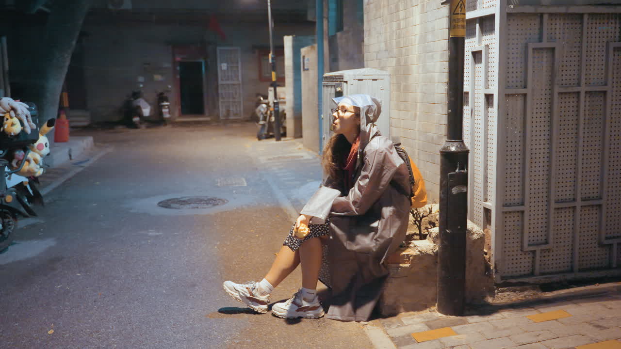 Mensajera descansando después de una entrega. Mensajera agotada encuentra un momento de paz. Una mensajera asiática se detiene contra una puerta de la ciudad bajo la lluvia. Escenario nocturno sombrío con una repartidora cansada en un callejón de la ciudad.