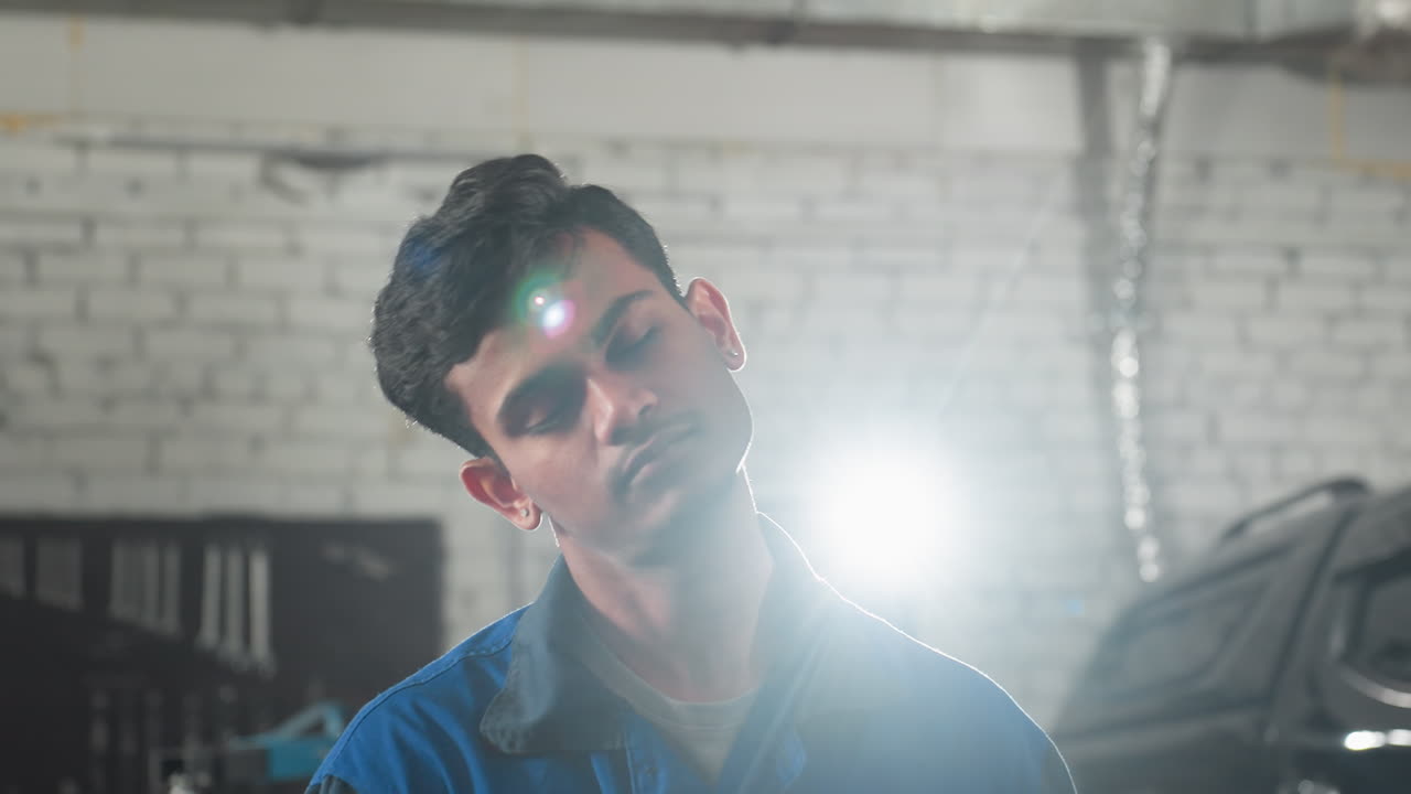 mecánico en uniforme azul inclinando la cabeza hacia la izquierda y la derecha con los ojos cerrados, iluminado por una luz de fondo brillante en un taller de automóviles, coche estacionado en el fondo