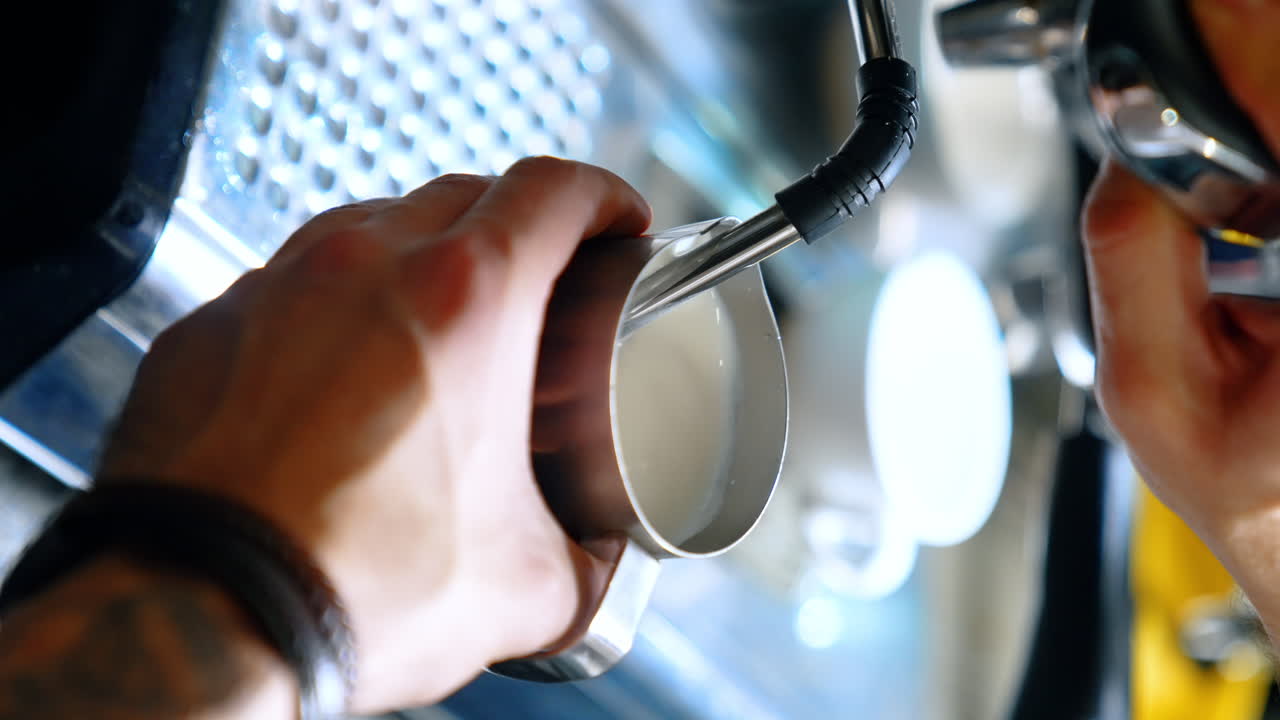 Barista's hands hold the metal pot filling it with hot milk from coffee machine. Making latte or cappuccino. Close up. Vertical view.