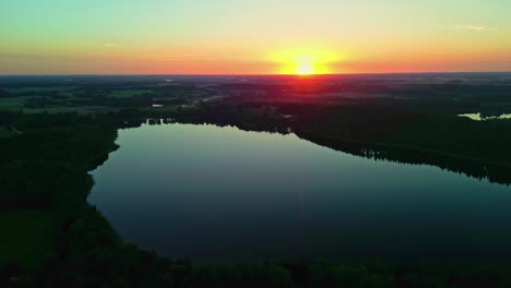 aerial shot flying backward revealing a serene lake at sunset surrounded by trees with vibrant sky colors reflected in the water