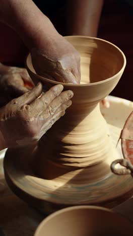 a close-up shot shows a skilled potter teaching and guiding a student's hands to shape a vase from wet clay on a spinning potter's wheel during an art class