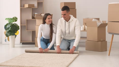 front view of a young couple in a new house sitting on the floor rolling a carpet 1