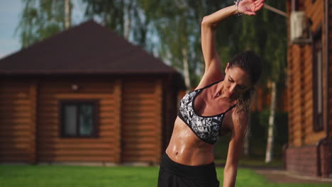 A-slim-and-beautiful-woman-in-sports-clothes-with-an-open-press-is-preparing-to-start-training-on-the-lawn-near-her-house-Performing-slopes-flexing-back-muscles