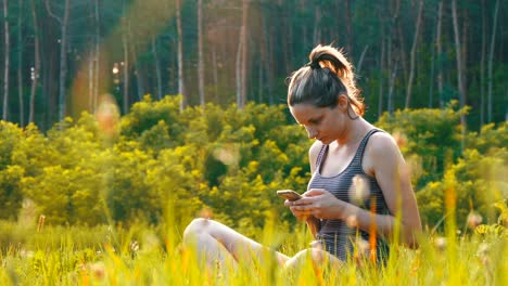 happy young woman sitting on green lawn and uses smartphone on scenic field at sunset background