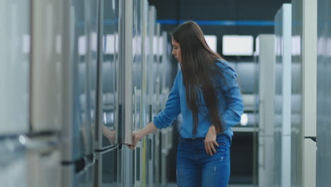 a young woman to open the refrigerator door to store appliances and compare with other models to buy the new house