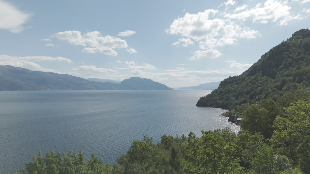 Rekaman drone gerak lambat yang menyingkap dari balik pepohonan melihat ke arah Fjord di Norwegia pada hari yang cerah dan berawan dengan pegunungan dan kehijauan di latar belakang serta air yang memantul LOG