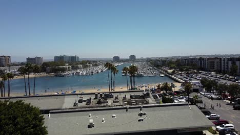 Aerial-drone-shot-ascends-over-a-busy-street-in-Marina-del-Rey-and-flies-towards-the-harbor-that-is-filled-with-boats-and-finally-looks-down-on-the-sandy-beach-and-the-people-enjoying-the-water