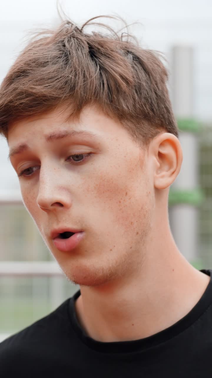 Young caucasian man with freckles, showing a pensive expression with open mouth, engaging in conversation or deep thought, standing outdoors against a soft background, vertical close up shot