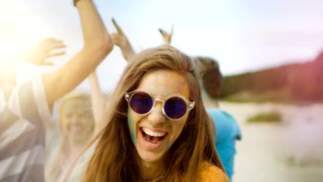 close-up portrait of a beautiful young girl with sunglasses standing in the crowd of people celebrating holi festival. people throwing colorful powder in her back. her face and clothes are covered with colorful powder.