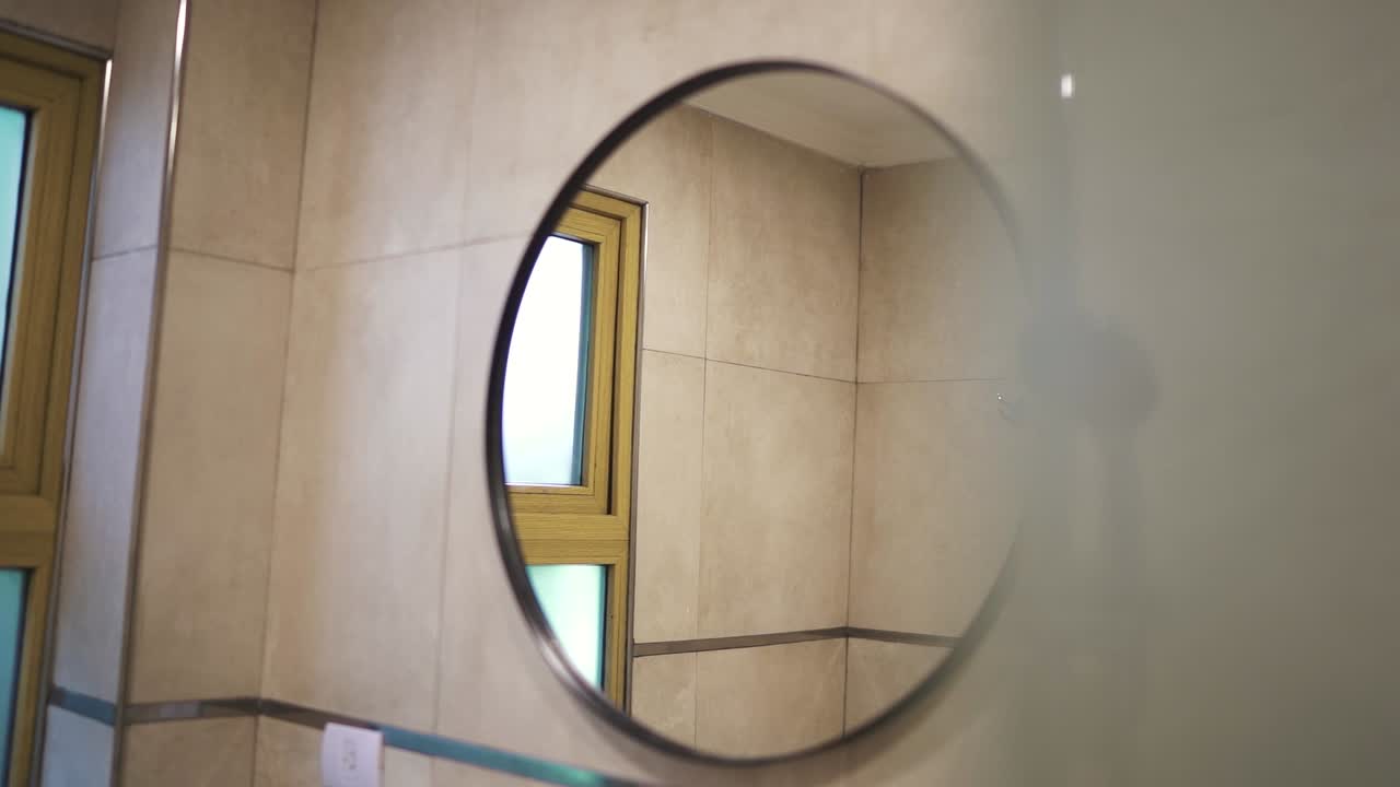 A slow-moving shot captures the round mirror in a luxury hotel room's bathroom, reflecting elegant decor and upscale amenities, showcasing a tranquil and stylish environment.