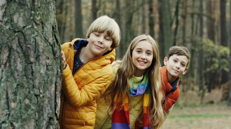 Portrait of two cute boys and one girl looking at camera behind a tree trunk in the forest