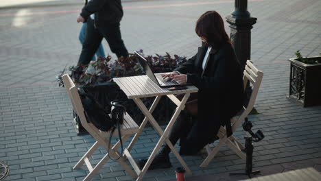 mujer profesional trabajando en una computadora portátil al aire libre en una cafetería rodeada de sillas de madera, plantas y trípode, sentada en una mesa con una taza de té, personas pasando en el fondo en una pasarela pavimentada