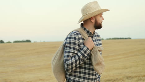 jeune agriculteur portant un chapeau marchant dans le champ avec un sac de grain et regardant les côtés