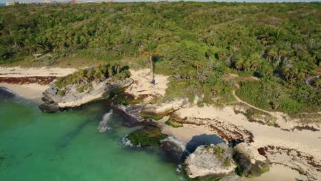 Vista-Aérea-Del-Agua-Azul-Chocando-Contra-Las-Rocas-En-Los-árboles-De-La-Playa-En-El-Fondo