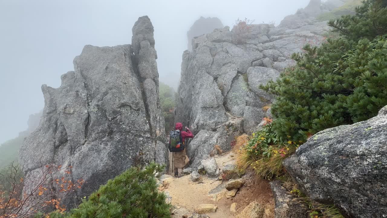 Capture of a backpacker on a narrow, misty trail between rocks in Nagano's Northern Alps, Japan. The scene conveys a sense of wanderlust, exploration, and overcoming nature's challenges