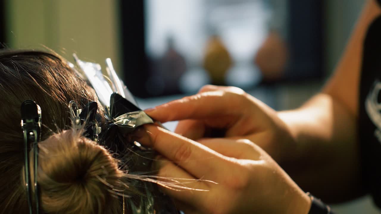 Stylist coloring hair of customer. Colorist places hair of client on foil and attaches clips as part of color treatment process. Salon owner and client receiving services. Close up.