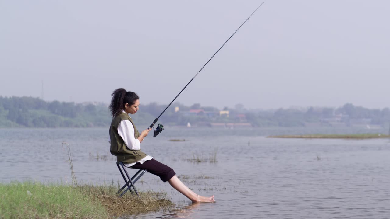 Seorang gadis Asia Selatan mengenakan rompi luar ruangan sedang duduk memancing di tepi danau yang tenang, bersantai dengan kakinya di air dangkal