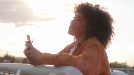 black woman wearing headphones and browsing phone while standing on the bridge