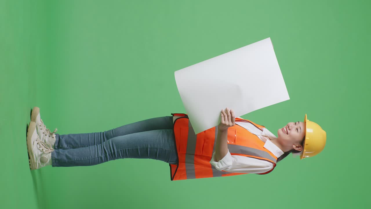 안전 헬을 착용한 아시아 여성 엔지니어의 몸 전체 측면 뷰, 그녀의 손에 청사진을 보고 녹색 스크린 배경 스튜디오에 서있는 동안 주위를 둘러보고 있습니다.