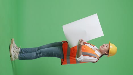 full body side view of asian female engineer with safety helmet looking at blueprint in her hands and looking around while standing in the green screen background studio