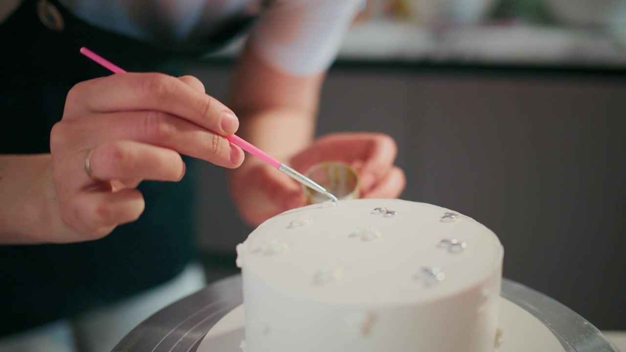 Baker carefully applies edible glitter to a white cake. Hands steady and focused, showing skillful artistry. Soft lighting highlights the delicate decoration process. Ideal for wedding, birthday