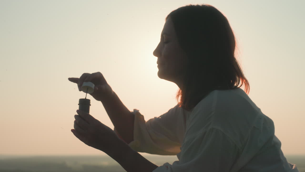 Close up profile of woman blowing soapy bubble wand into warm sunset light, silhouette illuminated by glowing sky, with gentle breeze moving hair and floating bubbles in tranquil outdoor moment